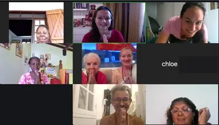 A six-tile video call grid of participants pressing palms together in namaskar, with a seventh tile showing the name label chloe. Top left, an older woman with short grey hair stands in a doorway with wooden slatted doors. Top centre, a smiling woman with long dark hair and a red bindi wears a red top in front of a shelf holding a small deity figure and framed pictures. Top right, a smiling young woman in a pink top. Middle left, an older woman in a pink floral top sits beside a KAILASA GUADELOUPE banner and a small shrine. Middle centre, two older women, one in red and one in peach, smile with palms pressed together. Bottom centre, a smiling man with grey hair, glasses and an orange-brown garment presses his palms together. Bottom right, a woman with curly dark hair and glasses touches her hair.
