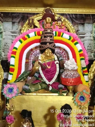 A seated golden deity murti of Annapoorana in a temple shrine, dressed in a magenta sari with a green and gold border, a gold necklace and a white flower garland. A tall black and gold crown-like headdress rises above her head, and her right hand is raised in abhaya mudra. A large rainbow-coloured flower arch in pink, yellow, red, white and green frames the figure. In front, a kalasha pot with white flowers rests on a brass stand, and small brass figures and floral decorations surround the golden throne. Behind the murti, a painted temple backdrop and gold framing are visible. The photo carries a timestamp of 15 Oct 2026 12:03:34 pm and the captions Periya Seeragapadi, Tamil Nadu and kailasa seeragapadi.