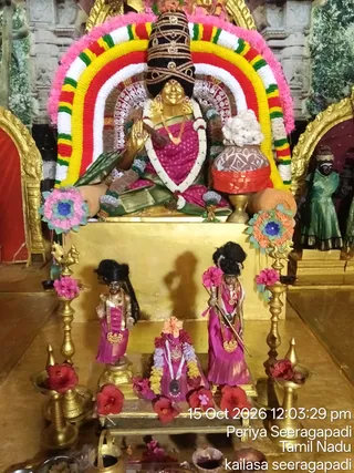 A photograph of a shrine altar with a golden deity murti of a seated goddess, adorned with a tall black and gold crown, a golden necklace and a pink and green sari. Her raised hand is a blessing gesture, and she sits before a red, yellow, white and green floral arch with pink garlands. A brass kalasha with white flowers stands at her right. Three smaller figures in pink and magenta garments, with gold necklaces, are placed below on a golden platform, with red hibiscus flowers scattered around and brass lamps on either side. A green-clad figure stands in a side niche at right. A timestamp and location caption, reading 15 Oct 2026 and Periya Seeragapadi, Tamil Nadu, appear in the lower right corner.