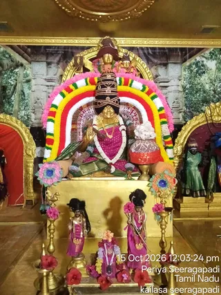 A photograph of a golden deity murti seated on a gold pedestal inside a temple shrine, decorated for darshanam. The seated goddess wears a magenta sari with a green-and-gold border, layered garlands and gold jewellery, and holds one hand raised in abhaya mudra. A tall dark crown-like headdress rises above her head, and an arch of red, yellow, white and green garlands frames her. Beside her stands a red-and-white ritual pot topped with white flowers. Two smaller magenta-clad figures stand in front holding a staff, with a small seated figure between them, all decorated with red hibiscus flowers. Brass oil lamps flank the pedestal, and a painted forest backdrop with carved stone pillars surrounds the shrine. A timestamp overlay reads 15 Oct 2026 12:03:24 pm.