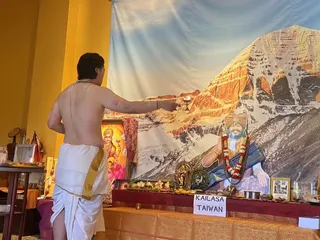 A bare-chested man in a white dhoti with a gold border stands with his back to the camera, extending his right arm to hold a lit oil lamp toward an altar. Behind the altar is a large backdrop of a snow-capped mountain face lit warm orange by sunlight. On the altar are a deity murti, brass offering vessels, flowers, fruit and a framed photo, with a portrait of a man in a saffron turban and a garland of red and white flowers and rudraksha beads. A white sign reads KAILASA TAIWAN. A picture of a goddess in red hangs at left beside a yellow wall. Setting suggests a Laghu Puja aarti in a devotional room.