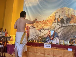 A shirtless man in white dhoti with gold border stands with his back to the camera, raising a small stick toward a shrine. The shrine has a mountain backdrop showing a snow-capped peak, and a large printed cutout of a figure in a blue garment with a turban, garlanded with red and white flowers, is set on a table with brass lamps, flowers, framed photographs and a red-and-gold cloth. A paper placard on the table reads KAILASA TAIWAN. A yellow wall and a Shabd-Ved book are visible at left.