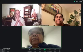A Zoom video call screenshot with three participant tiles. Top left, a woman with glasses and a white top sits with palms pressed together in namaste in a dining room; behind her a figure in orange and gold robes with a decorated ceremonial collar sits on a table near offerings and a framed deity picture. Top right, a woman with dark hair and a tilak on her forehead, wearing a dark top, sits before a framed painting and a monstera plant. Bottom centre, an older woman with glasses and a dark blue jacket is shown in close-up. The tile labels read marth, Shanthi Ravishankar and Indrani S, and the toolbar shows Participants 3.