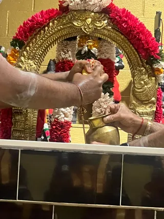 Close photo of a linga abhishekam at a shrine decorated with gold arch frame and red, white and orange flower garlands. Two hands, one bare-armed with white sacred ash smeared on the forearm and a red thread on the wrist, pour or press an offering over a dark stone linga. Beside them a brass kalasha/vessel and a small brass trident stand on the shrine ledge. Only partial arms and hands are visible; no full person's face is shown.