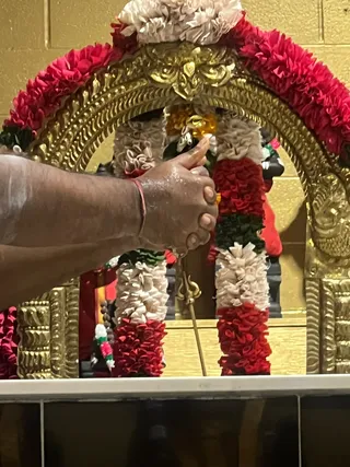 A close photograph of a deity shrine framed by an ornate gold arch decorated with red, white and green flower garlands. Only a partial arm and hand of a person is visible at left, the hand raised near the garlands with a thin wooden stick or rod pointing downward, apparently performing a ritual near the deity murti. A gold trident emblem is visible on the stand, and a dark murti sits behind the garlands. No full face or other physical people are visible.