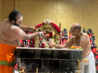 A photograph of a shrine ritual inside a room with yellow-gold brick walls. On the left, a bare-chested priest in an orange dhoti with green borders pours liquid from a brass vessel, with a white tilak smeared across his forehead and chest. On the right, an older bald priest in a white dhoti and bare chest, wearing a rudraksha mala, holds a brass plate with orange fruit. Behind them, a gold arch is draped with red and white flower garlands, and rows of black deity murtis wear red garments and flower garlands. A brass oil lamp and a small brass figure sit on the black-tiled altar, indicating abhishekam and puja.