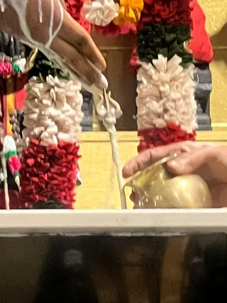 Close-up photo of a ritual abhishekam in progress. A hand pours a thin white milky stream from a golden vessel over a deity murti or linga, partly visible at the top left. Garlands of white and red flowers with green leaves hang over the figure, and a trident-like ornament is visible at the centre. A golden wall and a white ledge form the background. Only a partial hand is visible, so no other people are clearly visible.