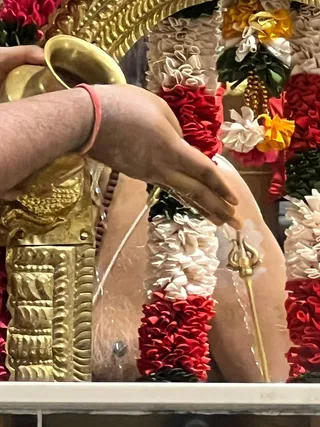 Close photograph of a ritual abhishekam on a deity murti. A hand holding a brass vessel pours liquid over the figure's bare torso, which is draped with red and white flower garlands. A gold trident-shaped ornament and a gold-embroidered garment appear at the left, with a golden arch of ornamentation above. A man's bare forearm with a red thread wristband is visible at the top left. The deity murti is not clearly identified, and no face is shown.