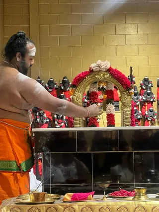 A shirtless priest with a bare, sweat-sheened back and a bun of dark hair stands at a shrine, lighting an oil lamp with a flame held near a gold arch decorated with red and white flower garlands. He wears an orange dhoti with a green and gold border, and white sacred ash marks on his forehead and arms. Behind him, rows of black stone deity murtis are garlanded with red and white flowers and draped in red cloth, set against a gold brick wall. On the altar in front are brass plates, a kalasha-style brass vessel, incense sticks with smoke rising, and a plate of red petals. The scene shows a puja in progress, with one visible person.