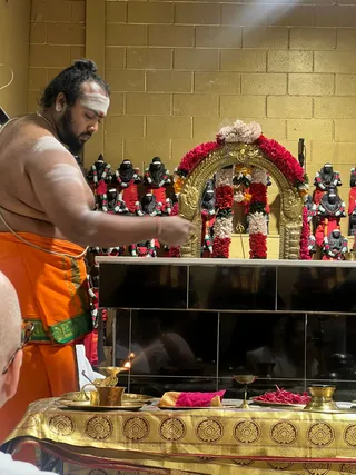 A bare-chested priest with a white ash band across his forehead and a red kumkum mark stands at a golden altar, wearing an orange dhoti with a green border and a sacred thread. He holds a ritual implement over an arch of red and white flower garlands framing a row of black stone deity murti with garlands and red cloth. Brass vessels, a lit oil lamp, a gold-bordered cloth and a plate of red flower petals sit on the altar. The setting is a yellow tiled wall, consistent with a puja or abhishekam being performed.