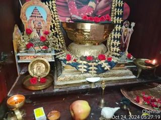 A photograph of a home shrine altar with no people visible. A large polished brass vessel with a gold-coloured base sits at the centre, draped with a navy blue cloth bordered in gold embroidery and decorated with red roses and dark rudraksha-like beads. Behind it hangs a printed photo of a figure in red and saffron clothing with a garland. A printed paper picture on the right shows a standing figure in an orange turban and a long rudraksha-style necklace holding a staff. On the left are small brass deity figures, a red-framed picture of a temple gopuram with a deity, and red roses scattered across the altar. Brass oil lamps with lit flames, a brass bell, a white conch shell, an apple, and a copper tray of red roses rest on the wooden shelf.