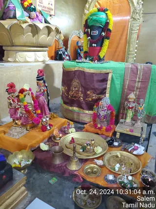 Photograph of a temple shrine interior with a dark Shiva lingam wearing a green cloth, a red-and-yellow flower garland and a horizontal tripundra mark on its front. In front, golden plates hold flowers, brass bells, a small oil lamp, bowls of offerings, a conch shell and a kalasha-style brass vessel on orange and maroon cloths. Several metal murtis are arranged in the foreground, including a pair of standing figures with flower garlands and red hibiscus flowers, a seated deity with a garland and a dancing or standing deity in green, along with a maroon cloth bearing printed deity figures and a bull. No people are visible. A timestamp reads Oct. 24, 2024 10:31:04 a.m. Salem, Tamil Nadu, and a sign reads KAILASA SALEM BHARATH.