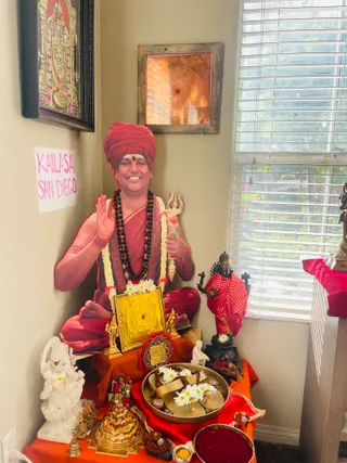 A corner altar in a room with cream walls and white window blinds. Seated at the centre is a life-size cutout figure of a smiling man in a red turban, red robes and a rudraksha mala, holding a trident and raising one hand in blessing; garlands of white flowers hang from his neck. Only a printed cutout is present, so no living person is shown. Before him sit a golden Sri Yantra plate, a brass tray of white chrysanthemums, kumkum and turmeric, and a white marble Ganesha statue on the left. A framed copper yantra and a framed painting of a deity hang on the walls, and a dark murti stands on a red cloth at right. A handwritten pink-and-white sign reads KAILASA SAN DIEGO.
