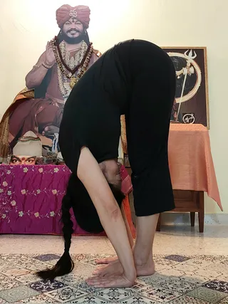 A woman with a long dark braid performs a standing forward bend in black athletic clothes on a patterned rug in a home room. Behind her, a large printed cutout of a smiling man in an orange-red turban, rudraksha mala and layered flower and gold necklaces, one hand raised in blessing, stands beside a framed trishul-and-circle image. A pink embroidered cloth covers a side table with a small photo and a red tilak-marked portrait, and a peach-coloured cloth covers another table at right.