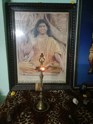 A framed printed portrait labelled "PARAMAHAMSA NITHYANANDA" hangs on a wall above a home shrine. The portrait shows a seated man in orange-saffron robes with a white beaded mala necklace, a red tilak on his forehead, and long swept-back dark hair, one hand raised in blessing against a backdrop of large banyan tree roots. In front of the portrait stands a brass oil lamp (kuthu vilakku) with a lit flame. The shrine table is draped with a leopard-print cloth and holds yellow marigold flowers, a small white shell, a glass shivalinga and a brass bell. A partly visible blue framed picture with Tamil text stands to the right. No people are present.