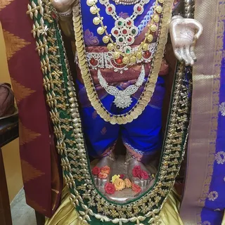 A close view of a deity murti. Its feet rest on a silver base decorated with red, orange and yellow roses and small silver cups. A burgundy cloth with gold triangular patterns and a blue patterned sari border frame the scene at the edges. No physical people are visible.
