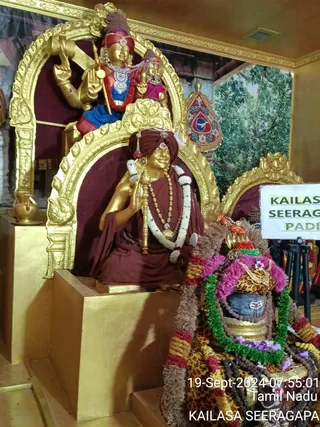 A golden statue of a seated guru in a maroon robe and maroon turban sits in a gilded arched niche, holding a staff in one hand and a rudraksha mala with a golden pendant around the neck. Above and behind, a golden deity murti in a red-and-blue garment with a crown and a smaller golden figure sits in a second niche. A brass kalasha stands at left. In the foreground a black stone Shivalinga is dressed in a leopard-print cloth, green and pink flower garlands, and rudraksha beads, with a placard reading KAILASA SEERAGAPADI beside it. A green tree backdrop and a red-and-gold emblem are visible. No living people are present.