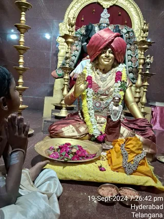 A photograph of a golden statue seated on a throne inside a temple-like hall with polished maroon stone floors and tall brass oil lamps. The statue wears a red turban, layered gold and jewelled necklaces, a flower garland of white, yellow and pink blooms, and a red-gold silk dhoti, with one hand raised in blessing. In front stand a brass tray of pink roses, two small bowls of kumkum, and a folded yellow-orange cloth with a jewelled ornament. On the left a child in white clothes sits with palms pressed together in prayer. Behind the statue a deity murti with a crown is visible. The date 19-Sept-2024 7:26:42 pm and Hyderabad Telangana are printed in the lower right corner.