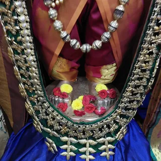 Close-up photograph of a deity murti seated in a green and gold embroidered alcove with a royal blue cloth beneath. The figure wears a maroon and gold silk garment with a silver bead necklace. In front, a shallow silver offering tray holds red roses and yellow marigold flowers arranged around a silver form. No physical people are visible; only the lower body and draped silk of the murti appear.