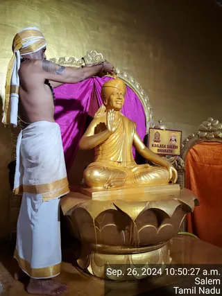 A photograph of a priest performing abhishekam on a golden statue seated in meditation pose on a lotus pedestal. The priest stands at left, bare-chested with a rudraksha mala, wearing a white dhoti with gold border and a white-and-gold headcloth, pouring liquid from a vessel onto the statue's head. The golden seated statue wears an orange-gold robe and a golden turban, one hand raised in blessing. Behind it hang magenta drapes, an ornate gold arch and an orange cloth. A small placard reads KAILASA SALEM BHARATH. A timestamp reads Sep. 26, 2024 10:50:27 a.m. Salem Tamil Nadu.