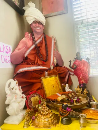 A photograph of a seated figure in a shrine corner with a white turban, a red-orange saffron-coloured robe with white-bordered shawl, and a long rudraksha mala around the neck, raising one hand in a blessing gesture and smiling. Around him is a ritual altar draped in yellow cloth with a white marble Ganesha statue, a golden Sri Yantra plate, a golden meru-shaped shrine piece, a brass tray holding rudraksha malas and flowers, a black Shivalinga with flowers and rudraksha, a red-robed dark deity murti and small gold idols. A handwritten pink sign reading partly LASA and DIEG is at left. The figure is in person and has a distinctive turban, robe and mala cues, so this is a probable Swamiji in person.