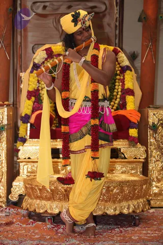 A photograph of a man standing barefoot on a red patterned carpet in front of an ornate golden throne draped with marigold and rose garlands. He wears a yellow turban decorated with a peacock-feather brooch and jewels, a yellow dhoti-style garment with a pink and green silk waist sash, a yellow shawl draped over one shoulder, multiple strands of white pearls, bangles and anklets. Red and orange flower garlands hang from his neck and he plays a wooden bamboo flute held to his lips. Peacock feathers appear on the golden pillars behind him. a person: the wearer's presentation has a turban, ornate saffron-yellow clothing and pearl and garland adornments, though the face is not clearly distinguishable, so the identity is uncertain.