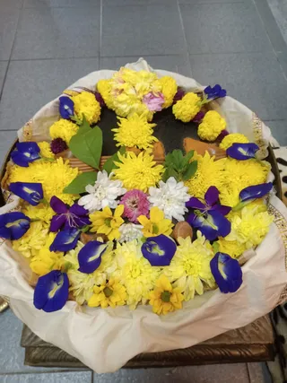 A photograph taken from above of a round offering arrangement on a tray draped with a white cloth edged in gold trim. Yellow and white chrysanthemums, yellow marigolds, a few pink and purple blooms, and blue-purple butterfly pea flowers are heaped around the rim, with green leaves tucked among them. A dark round surface sits in the centre, surrounded by small terracotta or wooden pieces. The tray rests on a grey tiled floor and a wooden stand. No people are visible.