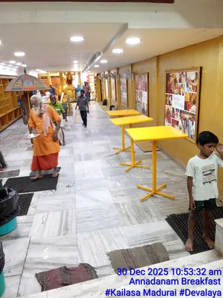 Interior corridor with polished marble floor and bright ceiling lights, lined with yellow tables and framed photo collages on a mustard wall. An elderly bearded man in orange robe and red dhoti, with a pink-orange shawl over his shoulder and a plate in hand, walks past a striped umbrella. A boy in a white t-shirt and dark shorts stands at right. Other people walk in the background near a golden statue. A framed collage labelled Darshan & Healing shows photographs of devotees and ceremonies. A timestamp overlay reads 30 Dec 2025 10:53:32 am, Annadanam Breakfast.