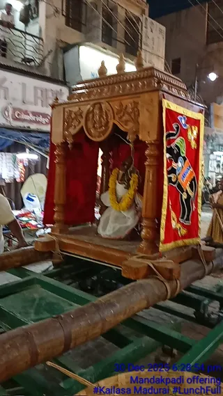 Night photograph of a wooden chariot or palanquin with carved wooden pillars and a carved deity panel above, mounted on long wooden poles and green-painted rails in a city street. Inside the shrine is a seated deity murti draped in white with a yellow marigold garland. A red hanging banner with gold border shows a painted black elephant with decorative trappings. Shop fronts, a balcony with a person standing, and hanging goods line the street. A blue timestamp overlay reads 30 Dec 2025 6:28:54 pm, Mandakpadi offering, #Kailasa Madurai #LunchFull.