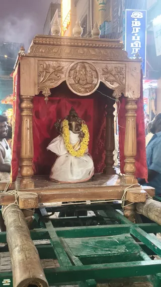 Night photograph of a wooden processional palanquin on a green wheeled cart in a street. The carved wooden canopy has a central medallion of Ganesha and floral carvings, with red cloth drapes. Inside sits a dark metal Ganesha murti wearing a white dhoti and a yellow marigold garland, seated on the red-draped platform. Wooden poles are lashed to the cart with rope. On the left, a smiling man in a white vest stands near the cart, and on the right a man in a blue shirt is partly visible among people in the crowd. Shop signs and lights line the street behind the procession.
