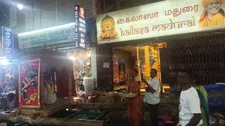 Night street scene in front of a shop with a large cream signboard reading கைலாசா மதுரை and kailasa madurai, with a golden deity emblem and a portrait of a smiling man in orange at the top right. A man in saffron robes, rudraksha mala and orange turban stands at center holding a brass tray of offerings, next to a wooden palanquin draped in red cloth with an elephant painting and a garland-decorated deity murti inside. A man in a white shirt holds a phone nearby, and a man in a white t-shirt and a woman in a green sari stand at right. Brass vessels and a gold-decorated doorway are visible inside the shop.