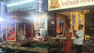 Night street scene outside a shop with a gold-framed entrance and signage reading "kailasa madu..." above. On the left, a wooden palanquin with a red cloth panel showing a painted elephant sits on bamboo poles, decorated with a white garland. In the center, a woman in saffron-orange sari and matching head wrap wears red and white beaded garlands and holds a woven tray of fruit and offerings. Beside her, a man in a white shirt and trousers holds up a phone to photograph her. Shelves of brass vessels and a red-and-gold deity image are visible in the background.