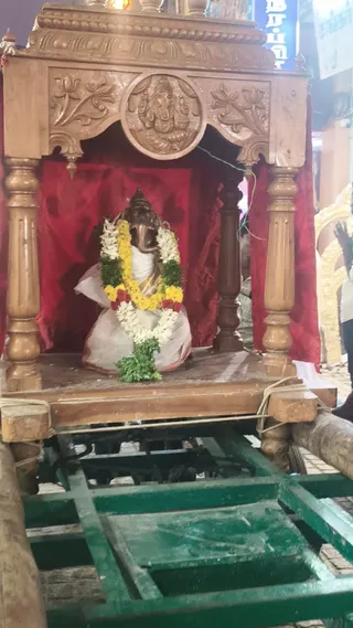 A photograph of a small wooden palanquin or chariot shrine on a green wheeled cart. Inside the carved wooden canopy, with a red cloth backdrop, sits a bronze Ganesha murti, adorned with a garland of white jasmine, yellow and orange flowers and green leaves, and draped in white cloth. The carved panel above shows a Ganesha relief flanked by lotus motifs. A few people's arms and legs are partly visible at the right edge, and a gold decorative arch appears beside the shrine. Stone pavement and a sign are visible in the background.