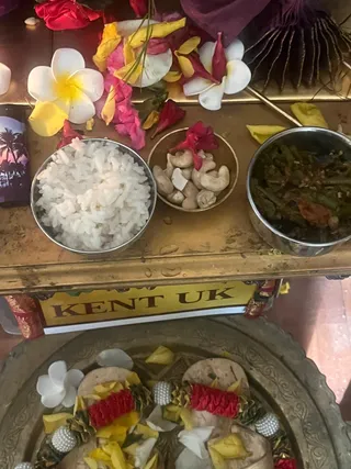 Overhead photo of a brass offering table at KAILASA Kent UK, decorated with scattered white plumeria and yellow and pink flower petals. Three metal bowls hold cooked white rice, cashew nuts with a red flower on a small brass cup, and a dark green vegetable dish. Below, a large ornate brass tray holds two small pieces of white offering material decorated with red garlands, yellow petals, white flowers and pearl-like beads. No people are visible.