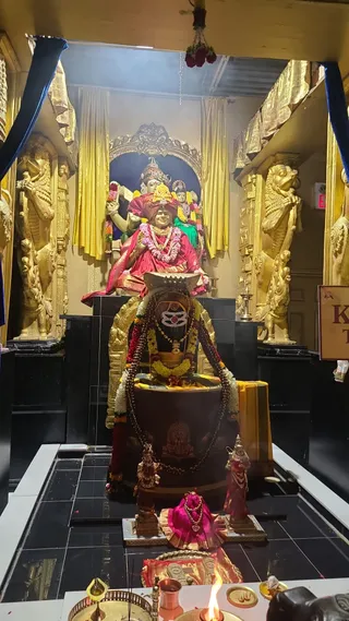 Photograph of a temple shrine interior with gilded lion-like guardian figures flanking a dark stone linga draped in garlands of yellow and orange marigolds, red roses and a rudraksha-bead mala. Behind it sits a seated golden statue in red and gold robes with a red turban and a garland of pink and white flowers, holding a mala in one hand, in front of a painted backdrop of multi-armed deity figures. Small figures in pink and red stand at the base of the linga, and in the foreground a lit oil lamp, a copper vessel, a brass plate and a flame sit on a black tiled floor. Gold drapes and blue cloth frame the scene. No people are visible.
