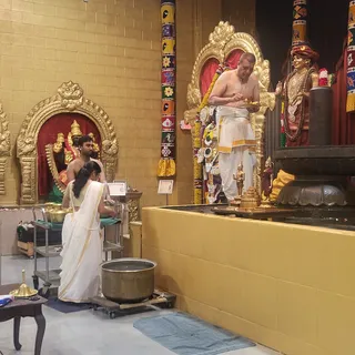 A photograph inside a gold-walled temple hall. In the centre, a bare-chested priest in white dhoti with a gold border stands at a raised stone abhishekam platform holding a small brass vessel, with a sacred thread across his chest. Beside him a tall deity statue in a red turban and gold necklaces stands near the platform. In the left background a deity murti sits in a golden arch with a red backdrop, and a bare-chested younger man in a white dhoti attends it beside a woman in a white sari with gold border who kneels at a steel cart holding brass vessels. A large bronze pot sits on a wheeled stand in the foreground, with a brass lamp on a small table at left. Garlands of marigolds hang on the arches.