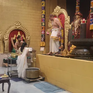 A temple interior with gold-toned walls and a gilded arched shrine niche holding a deity murti under a red cloth backdrop. A shirtless man in a white dhoti with gold border stands on a raised platform beside a large dark stone pedestal and a standing statue wearing a red turban, gold necklaces and rudraksha beads. Below, a man with bare upper body sits near a brass vessel, while a woman in a white sari with gold border kneels beside a stainless steel trolley holding brass pots. A large brass vessel sits on a wheeled cart, and a brass lamp rests on a small wooden table. Colourful striped pillars with decorative panels frame the scene. Ritual ablution activity is visible.