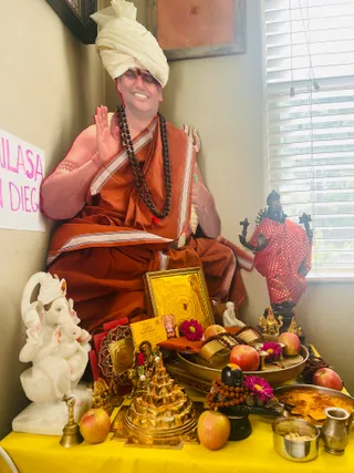 A large seated figure in a white turban and orange-brown saffron robes with dark rudraksha mala beads raises one hand in blessing beside a shrine corner. The figure may be a statue or life-size depiction, with a smiling face and a red-orange tilak. In front lies a yellow-draped altar with a white marble Ganesha, a golden shri yantra and meru, a gold-framed image, a red deity murti, apples, pink flowers, brass dishes, a black shivalinga with rudraksha mala, and a small brass kalasha. A pink handwritten sign reading 'KAILASA SAN DIEGO' is on the left wall, and a 'GURU PURNIMA' card sits on the altar. Sunlit window blinds are at right.