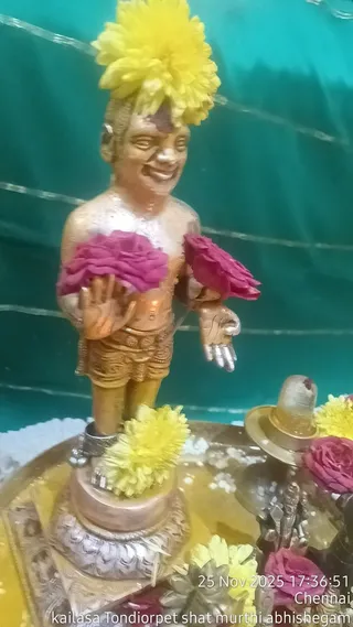 Close photo of a small golden statue of a standing deity murti, bare-chested and wearing a patterned lower garment, with a yellow flower crown and pink roses held in its hands. It stands on a silver pedestal beside a small shivalinga with yellow and pink flowers scattered around, on a golden tray with a turquoise backdrop. The scene reads as a shat murti abhishekam ritual, with a timestamp overlay of 25 Nov 2025 17:36:51 Chennai. No physical people are visible.