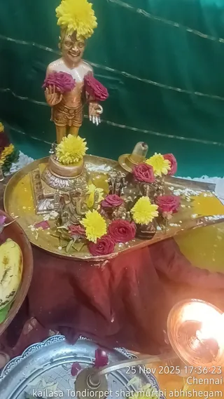 A photograph of a ritual arrangement on a golden tray draped with a maroon cloth, set against a green backdrop. A small golden-brown standing figure of a child-like deity with a yellow flower crown and red flowers draped over the arms stands at the back. Around its base are a silver-toned Ganesha figure, small metal deity figurines, a brass shivalinga-style vessel, yellow chrysanthemums and red roses, with scattered turmeric and rice. A brass oil lamp burns at right and a banana sits at left. No people are visible; the scene is a puja arrangement of deity murtis and offerings.