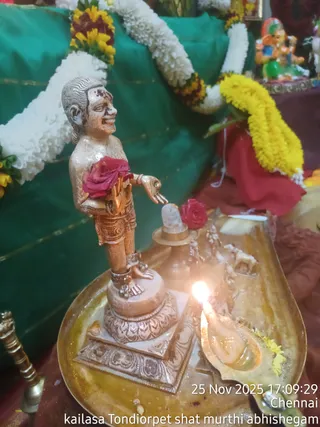 A photograph of a small metallic figure of a standing devotee-like statue, golden-bronze in colour and bare-chested with a wrapped lower garment and anklets, holding a red rose against its chest and extending one hand toward a small white shivalinga on a brass pedestal. A lit oil lamp flame sits on a brass tray in the foreground, with a yellow flower beside it. Behind the figure are white and yellow marigold garlands draped over a green cloth backdrop, and a small multicoloured deity figure stands on a shelf at the upper right. A timestamp overlay reads 25 Nov 2025 17:09:29 Chennai and a caption reads kailasa Tondiorpet shat murthi abhishegam. The figure is a statue, not a physical person.