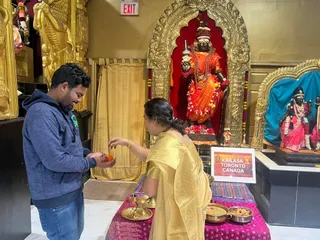 A photograph inside a temple shrine room with gold-finished arches and an EXIT sign above. A woman in a gold and beige sari stands at a table draped in a pink and purple cloth, offering a fruit or sweet to a man in a blue hooded sweatshirt and jeans who stands at left and holds an orange. Behind them a dark stone deity murti in an orange and saffron garment, with rudraksha malas, flower garlands and a crown-like headdress, stands in a red-backed gold niche. To the right another deity murti in a pink sari and garlands sits on a black pedestal beside a blue backdrop. On the table are brass plates, a brass lamp, and bowls of food. A white sign reads KAILASA TORONTO CANADA.