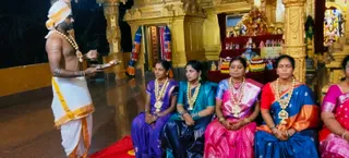 A priest in a white dhoti with orange-gold borders and a white head cloth stands at left, holding a brass plate and a small lamp while speaking, his bare chest marked with white and yellow paste and a beaded necklace around his neck. Five women in silk saris sit on a red mat in front of a golden temple structure: one in purple, one in teal and navy, one in pink with a gold-bordered sari, one in blue with orange borders, and a partly visible woman in pink at right. Each wears gold necklaces and bangles. Behind them is an altar with a small banner reading KAILASA HYDERABAD, decorated deity figures and flower garlands, and colourful cloth hangings on the pillars. This appears to be a puja ritual with women seated for the offering.
