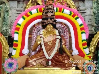 A golden deity murti of Goddess Kamakshi Amman seated in a lotus-style posture on a golden base, dressed in a rust-brown sari with gold dot pattern. She wears a tall black crown-like headdress with gold ornaments, a white and gold garland, and gold jewellery. Behind her is an arch of red, yellow, green, white and pink flower garlands framing the shrine. Pink and blue floral decorations sit at the lower corners, and the setting is a decorated temple altar with gilded surroundings. A timestamp in the corner reads 13 Oct 2026 11:55:59 am, Periya Seeragapadi, Tamil Nadu.