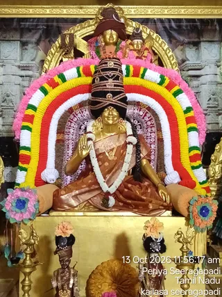 A seated golden deity murti of Goddess Kamakshi Amman, adorned in a rust-brown silk sari with gold border, a long white flower garland, gold jewellery and a tall black crown with gold ornaments. The murti raises one hand in abhaya gesture under an arch of pink, white, yellow, red and green flower garlands. Floral decorations in pink and orange sit at the sides, and small brass oil lamps flank the base. The photo carries a timestamp of 13 Oct 2026 and a Periya Seeragapadi, Tamil Nadu, kailasa seeragapadi caption.