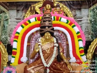 A golden deity murti of a seated goddess, adorned for alankaram, sits in a shrine. She wears a brown and gold patterned sari, a gold necklace and earrings, and a tall black and gold headdress. Her right hand is raised in abhaya mudra. A large arch of garlands in yellow, red, green, white and pink frames her, with pink flowers above. A painted temple backdrop with carved pillars and greenery stands behind the murti. No people are visible.