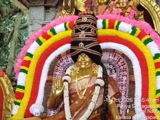 A golden murti of a goddess seated in a temple shrine, adorned with a black crown banded in gold and jewelled ornaments, a gold necklace, and a white flower garland. She wears a rust-brown sari with a gold-patterned border and raises one golden hand in abhaya mudra. Behind her a large arch of concentric garlands in pink, yellow, red, green and white frames the murti, and the timestamp reads 13 Oct 2026 11:55:41 am with the location Periya Seeragapadi, Tamil Nadu. Gold-coloured decorations and other murtis are visible at the top edge of the frame.