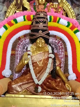 A golden deity murti of a goddess seated on a throne, decorated for darshan. She wears a dark cylindrical crown with gold bands and a gold ornament, a red-brown silk sari with gold motifs, a gold necklace and earrings, and a long white and green garland. Her raised right hand shows a gold palm ornament. Behind her is a rainbow-like arch of red, yellow, green and white flower strands, with a silver-patterned backdrop and gold arches above. A timestamp and location caption are printed in the lower right corner.