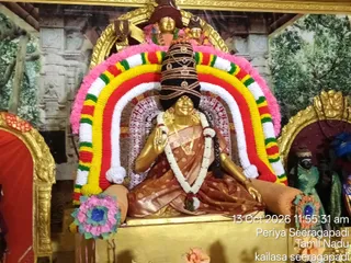 A photograph of a golden deity murti of Goddess Kamakshi Amman seated in a temple shrine, decorated for darshan. The goddess wears a tall black crown-like headdress banded with gold, a brown and gold sari, a white flower garland, and gold necklaces and earrings. Her raised hand holds a small object. A large arch behind her is framed by bright garlands in pink, yellow, red, green and white, with flower decorations at the base. Gold-framed arches and a painted backdrop of trees and temple carvings surround the murti. Statues of attendant figures stand at the right edge. A timestamp and location caption are printed over the lower right corner.