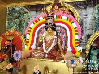 A photograph of a golden deity murti seated in a shrine decorated with a multicolour garland arch in red, yellow, white, green and pink. The central figure wears a dark tiered crown with gold ornaments, a reddish-brown sari with white dots, a white flower garland and a gold necklace, and raises one hand in blessing. Flanking shrine figures and fresh flower decorations in pink, orange and blue sit at the base, with a painted forest backdrop behind. No people are visible. A timestamp reads 13 Oct 2026 11:55:38 am, Periya Seeragapadi, Tamil Nadu, kailasa seeragapadi.